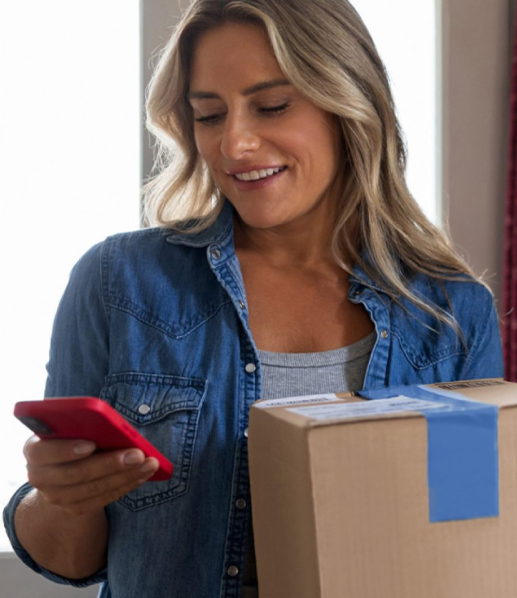 A smiling woman looks at her smart phone as she holds a package.