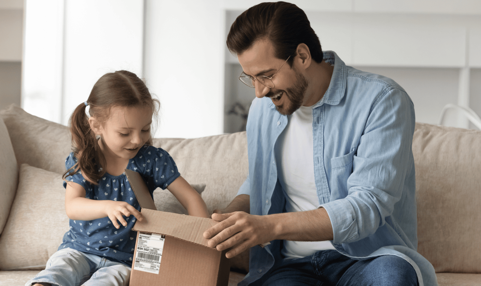 Adult and child sitting on a couch opening a small cardboard package together in a bright living room.